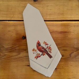 May include: A stack of beige cloth napkins with a decorative cardinal bird and autumn leaves design. The cardinal is red with a black mask and is perched on a branch with red berries and orange leaves. The napkins have a decorative stitched edge.