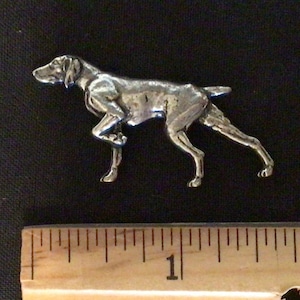 May include: A silver-colored figurine of a German Shorthaired Pointer dog in a running pose. The figurine is about 1 inch tall and is shown next to a ruler for scale.