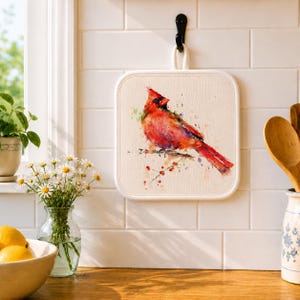 May include: A square, off-white pot holder with a watercolor painting of a red cardinal bird perched on a branch. The bird has red, orange, and black feathers. The pot holder has a white border and a loop for hanging.