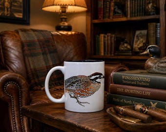 May include: A white ceramic mug featuring a detailed illustration of a bobwhite quail with brown, black, and white plumage. The mug has a solid dark blue interior and a curved handle, standing on a wooden surface next to stacked books and cigars.