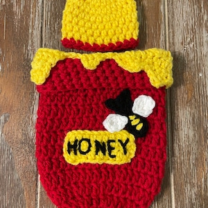 May include: A crocheted honey pot costume for a baby, featuring a red honey pot with a yellow top, a bee applique, and the word "Honey" on the front. The costume comes with a matching yellow crocheted hat with bear ears.