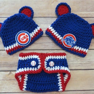 May include: A blue and red crocheted baby hat with bear ears and a Chicago Cubs logo. A matching blue and red crocheted diaper cover with baseball buttons.