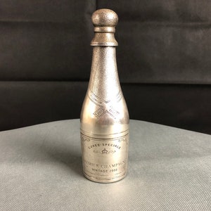 Peut inclure: Un objet argenté en forme de bouteille avec un dessus arrondi et une surface texturée. La bouteille porte l'inscription "Cuvee Speciale Premier Champagne Vintage 2000 France". La bouteille est posée sur une surface grise.