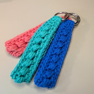 May include: Three handmade crochet keychains in pink, teal, and blue. Each keychain features a textured, bubble-like stitch pattern and a silver-colored metal ring for attaching keys. The keychains are displayed on a neutral-toned surface.