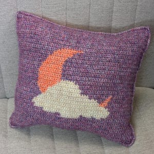 Könnte beinhalten: Ein quadratisches, gehäkeltes Kissen in Lilatönen mit einem Design aus einer orangefarbenen Mondsichel und einer weißen Wolke. Das Kissen liegt auf einer neutralen Oberfläche und zeigt die handgefertigte Textur und Farbpalette.