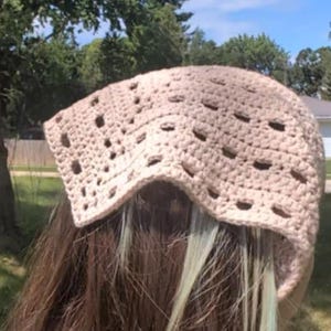 May include: A beige crocheted hat with a square shape and openwork design. The hat is worn over brown hair with streaks of light green. The background features green trees and a blue sky.