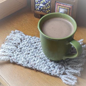 May include: A green mug filled with a warm beverage sits on a handmade, fringed coaster. The coaster is a light gray with flecks of color, and the mug has a simple, rounded shape. The scene is set on a wooden surface.