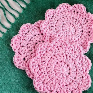 May include: Three handmade pink crochet coasters with a scalloped edge are displayed on a green textile. The coasters are made of a thick yarn, and the stitches are clearly visible. The coasters are arranged in a slightly overlapping pattern.