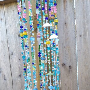 Puede incluir: Un carillón de viento de cuentas con hebras de cuentas coloridas en tonos de azul, verde, amarillo y rosa. Las hebras están adornadas con acentos de conchas marinas y vidrio, creando un tema costero.