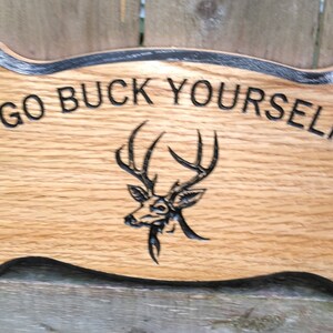 Puede incluir: Cartel de madera con la frase "GO BUCK YOURSELF" tallada en la madera. Una cabeza de ciervo está grabada debajo del texto. El cartel tiene un borde negro y un acabado natural de vetas de madera.