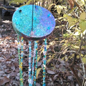 Puede incluir: Un móvil de viento hecho a mano con una parte superior redonda, brillante, azul y morada. El móvil tiene hilos de cuentas de colores en tonos azules, verdes y amarillos. El móvil de viento cuelga de una cuerda negra en un entorno natural al aire libre.