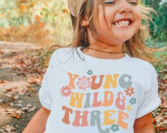 May include: A white t-shirt with a colorful retro design that says "Young Wild & Three" with flowers and a peace sign.