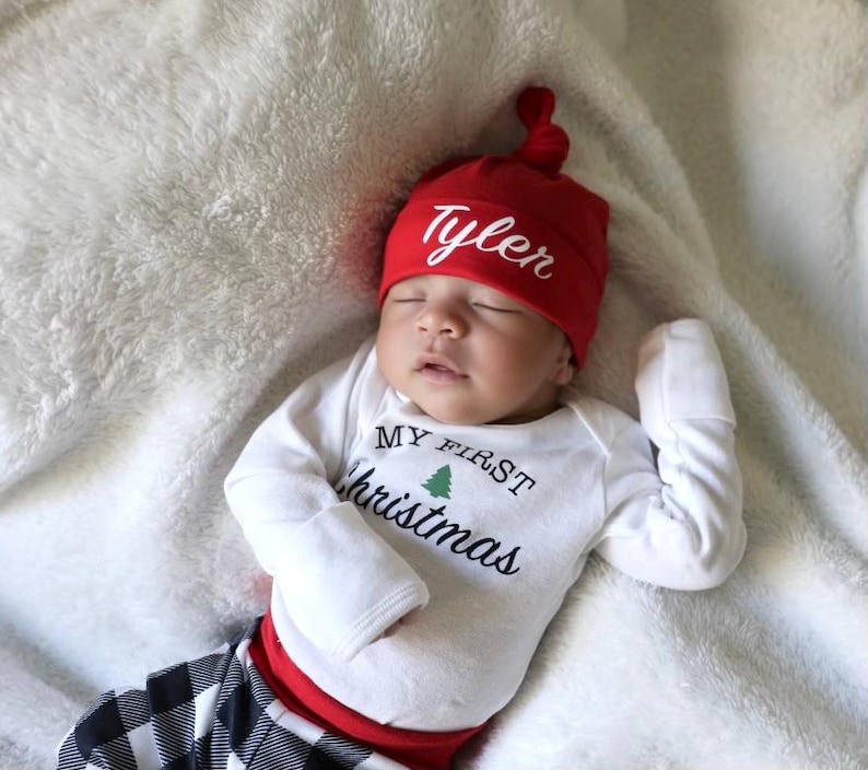 May include: A baby wearing a red and white Christmas outfit with a personalised knotted hat that says "Tyler" and a white onesie that says "My First Christmas".