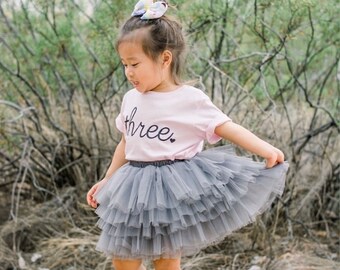 May include: A young girl wearing a pink t-shirt with the word "three" on it and a gray tulle skirt. She is standing in front of a green bush.