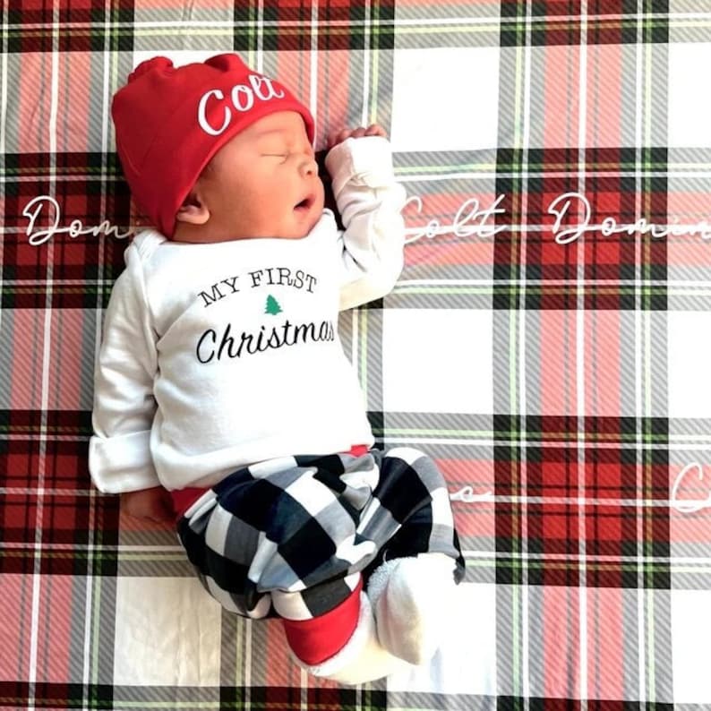 May include: A baby wearing a white onesie with the text "My First Christmas" and a green Christmas tree graphic. The baby is wearing a red and white plaid hat with the name "Coll" embroidered on it. The baby is lying on a red and white plaid blanket.