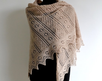 May include: A beige knitted shawl with an intricate openwork design and a scalloped edge. The shawl is displayed on a black mannequin, highlighting its texture and pattern. The neutral colour makes it a versatile accessory.
