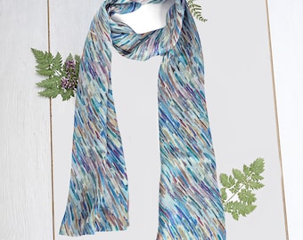 May include: A blue and white silk scarf with a watercolor-like pattern of diagonal lines. The scarf is folded in half and draped over a white wooden surface.