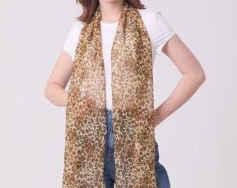 May include: A sheer, rectangular scarf with a leopard print pattern in shades of brown and tan. The scarf is draped over the shoulders. The model is wearing a white t-shirt and blue jeans.