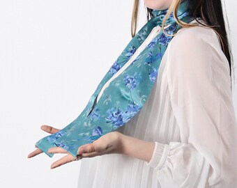 May include: A teal blue silk scarf with a floral pattern in shades of blue. The scarf is tied around the neck of a person wearing a white blouse.