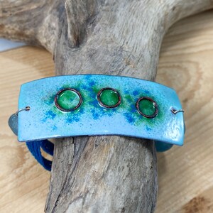 May include: A blue enamel bracelet with four green circles outlined in copper. The bracelet is on a wooden surface.
