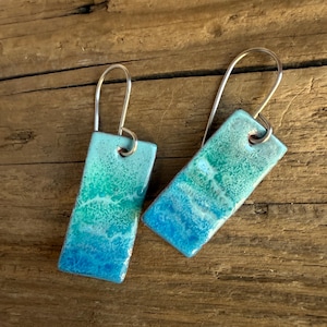 May include: A pair of rectangular earrings with a gradient design. The earrings transition from a deep blue at the bottom to turquoise and green at the top. They have silver-colored ear wires and a glossy finish, evoking a coastal theme.