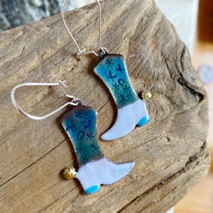 May include: A pair of silver dangle earrings with blue and white enamel cowboy boot charms. The boots have a white base with blue and green enamel designs and the text "HOT" on them.