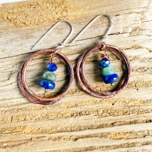 May include: A pair of dangle earrings featuring hammered copper hoops suspended from silver-toned ear wires. Each earring includes a vertical stack of three small beads in alternating shades of deep blue and pale green, secured with copper wire.