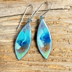 May include: A pair of artisan earrings with a teardrop shape. The earrings feature a vibrant, abstract design in shades of turquoise, blue, and brown, with a black outline. The ear wires are silver-colored. The earrings are displayed on a rustic wooden surface.