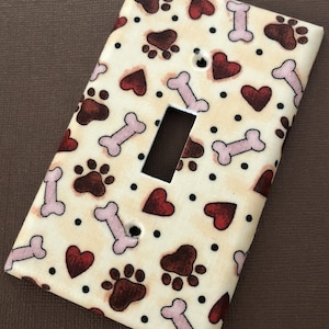 May include: A light switch cover with a cream background and a pattern of red hearts, brown paw prints, and pink bones.