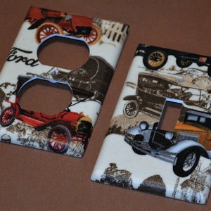 May include: Two vintage-style light switch and outlet covers featuring a white background with a pattern of antique cars. The outlet cover has two outlets and the switch cover has a single toggle switch. The text "Ford" is visible on the outlet cover.