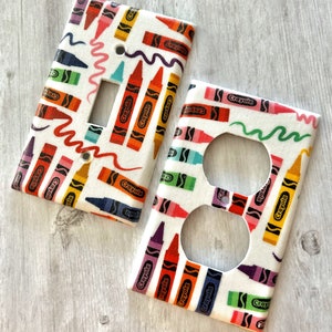 May include: Two white light switch and outlet covers with a colorful crayon pattern. The covers feature the word "Crayola" repeated throughout the design.