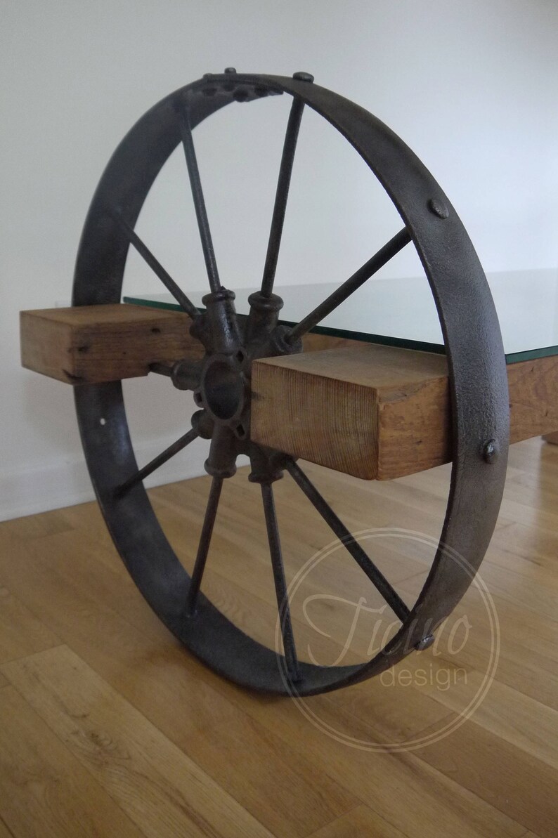Pode incluir: Uma mesa de centro r&uacute;stica feita de uma roda de carro&ccedil;a reutilizada, t&aacute;buas de madeira e um tampo de vidro. A roda &eacute; pintada de preto e tem um acabamento desgastado. As t&aacute;buas de madeira s&atilde;o tingidas de uma cor marrom natural. O tampo de vidro &eacute; transparente e adiciona um toque moderno &agrave; mesa.