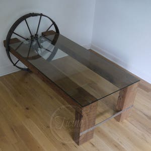 Pode incluir: Uma mesa de centro r&uacute;stica feita de madeira recuperada com tampo de vidro e base de roda de metal. A mesa tem um acabamento de madeira natural e &eacute; sustentada por duas pernas grossas de madeira.