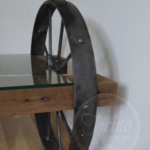 Pode incluir: Uma mesa de centro r&uacute;stica com tampo de vidro e base de roda de metal. A roda &eacute; pintada de preto e tem um acabamento desgastado. A mesa &eacute; feita de madeira recuperada e tem um acabamento de madeira natural.