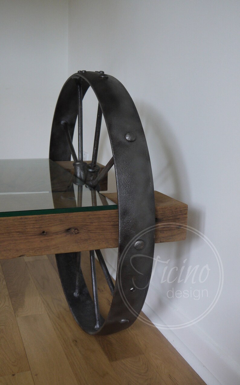 Pode incluir: Uma mesa de centro r&uacute;stica com tampo de vidro e base de roda de metal. A roda &eacute; pintada de preto e tem um acabamento desgastado. A mesa &eacute; feita de madeira recuperada e tem um acabamento de madeira natural.