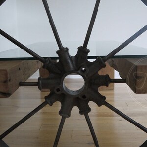 Pode incluir: Uma mesa de centro r&uacute;stica feita de uma roda de carro&ccedil;a reutilizada com tampo de vidro e apoios de madeira. A roda &eacute; de metal e tem um acabamento preto. A mesa &eacute; suportada por duas vigas de madeira.
