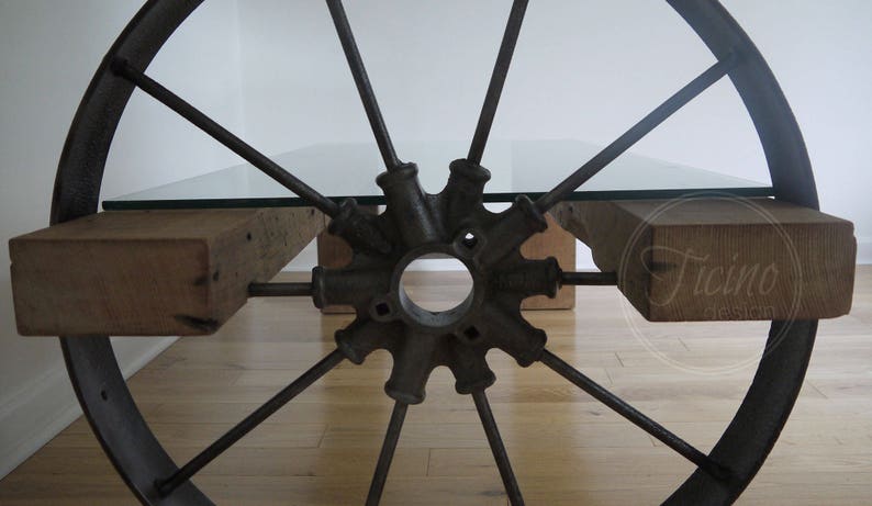 Pode incluir: Uma mesa de centro r&uacute;stica feita de uma roda de carro&ccedil;a reutilizada com tampo de vidro e apoios de madeira. A roda &eacute; de metal e tem um acabamento preto. A mesa &eacute; suportada por duas vigas de madeira.