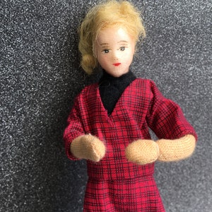 May include: A doll with blonde hair wearing a red and black plaid dress with a black turtleneck underneath. The doll has a painted face with red lips and brown eyes.