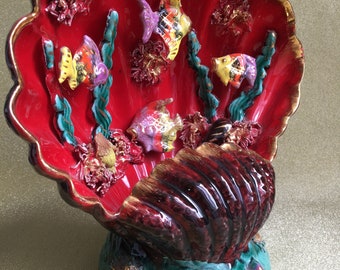 Può includere: Una conchiglia di ceramica rossa e marrone con una base verde. La conchiglia è decorata con pesci e coralli colorati.