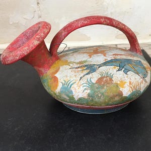 Peut inclure: Une cruche en céramique rouge et blanche avec une anse et un bec verseur. La cruche est décorée d'un poisson bleu et de motifs floraux verts et jaunes.
