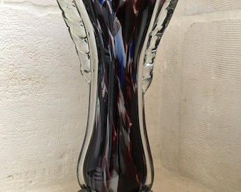 May include: A tall, clear glass vase with a flared opening and a wide base. The vase is decorated with swirls of white, blue, and red glass.