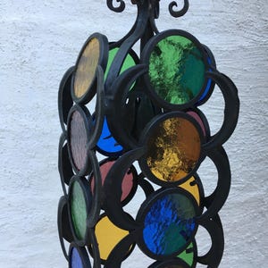 Può includere: Un'elegante lampada a sospensione con una struttura in metallo nero. Presenta una serie di pannelli circolari in vetro colorato nei toni del giallo, verde, blu e rosso. Il design è intricato e decorativo.
