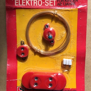Puede incluir: Un antiguo Elektro-Set para niños a partir de 7 años, empaquetado en cartón rojo y amarillo. El conjunto incluye componentes de plástico rojo, alambre de cobre y un conector blanco. El paquete tiene texto en alemán.