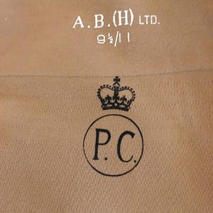 Può includere: Una superficie in tessuto color beige con testo stampato in bianco che recita A.B.(H) LTD. e 9 1/2 / 11. Sotto, un timbro a inchiostro nero mostra un simbolo di corona sopra le lettere P.C. racchiuse all'interno di un bordo circolare.