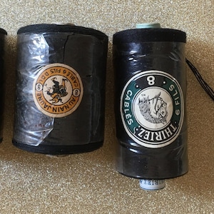 Peut inclure: Trois bobines de fil noir avec des étiquettes. Les étiquettes présentent du texte et des illustrations en jaune, blanc et vert. Une bobine a un cordon noir attaché. Les bobines sont enveloppées dans du plastique transparent.