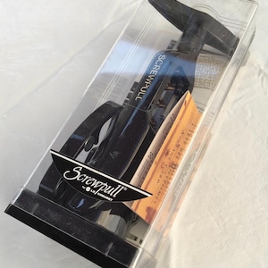 May include: A black and silver Screwpull wine opener in a clear plastic box. The box has the words "Screwpull by Le Creuset" printed on it.