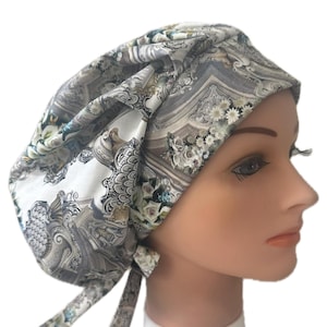 May include: A patterned surgical scrub cap featuring a floral and architectural design in shades of gray, white, and touches of blue and green. The cap has a tie closure at the back and is designed to cover the head.