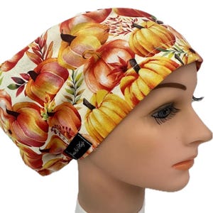 May include: A fall-themed scrub cap featuring a repeating pattern of pumpkins in shades of orange, yellow, and red. The cap has a white background with green and brown leaves. A small black tag is sewn on the side.