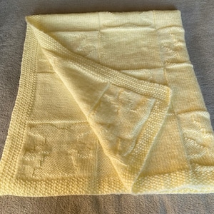 May include: A pale yellow knitted baby blanket with a square pattern. The blanket is folded in half and has a soft, textured surface.