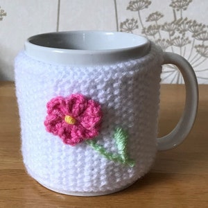 May include: White crocheted mug cozy with a pink flower and green stem. The cozy is on a white ceramic mug.